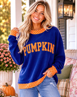 A woman wearing a royal blue crewneck sweatshirt with 'PUMPKIN' printed across the front in orange letters with white polka dots, paired with blue jeans, standing on a decorated porch with pumpkins and flowers.