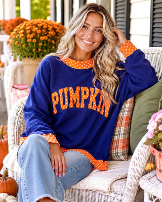 A woman sitting on a white wicker porch chair wearing a navy blue sweatshirt with orange polka-dot trim and 'PUMPKIN' written across the front in orange letters with white polka dots. She is wearing light blue jeans, with pumpkins, mums, and a house porch background.