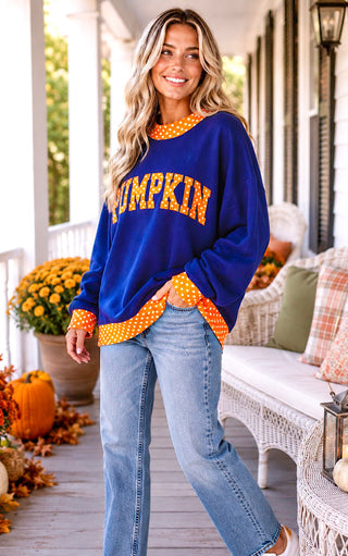 A woman stands on a porch wearing a royal blue long-sleeve sweatshirt with orange and white polka-dot trim and 'PUMPKIN' written across the front in orange polka-dot letters, paired with light blue jeans.