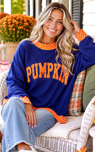 A woman sitting on a white wicker porch chair wearing a navy blue crewneck sweatshirt with 'PUMPKIN' written in orange arched letters with white polka dots, and matching orange polka dot cuffs and hem.