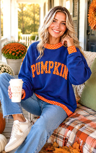 A woman sitting on a porch wearing a royal blue crewneck sweatshirt with 'PUMPKIN' written across the chest in orange letters with white polka dots. She is wearing blue jeans and white sneakers and holding a white coffee cup.