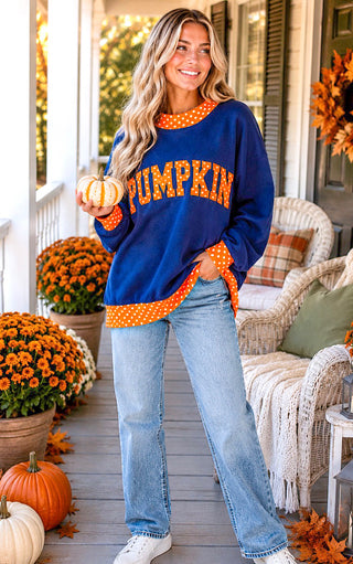 A woman stands on a decorated front porch wearing an oversized navy blue sweatshirt with orange polka-dot trim and 'PUMPKIN' written across the front in orange letters, paired with light blue jeans and white sneakers.