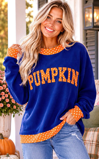 A woman wearing a royal blue long-sleeve sweatshirt with orange and white polka-dot trim and 'PUMPKIN' printed across the front in orange letters, paired with blue jeans in an outdoor porch setting.