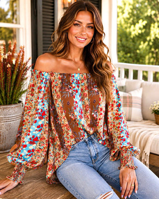 A woman wearing a colorful off-the-shoulder floral blouse with sheer mesh panels and long ruffled sleeves, paired with light-wash blue jeans, sitting on a porch.