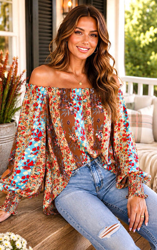 A woman sitting on a porch wearing a brown floral off-the-shoulder bohemian floral top with long ruffled sleeves and light-wash distressed jeans.