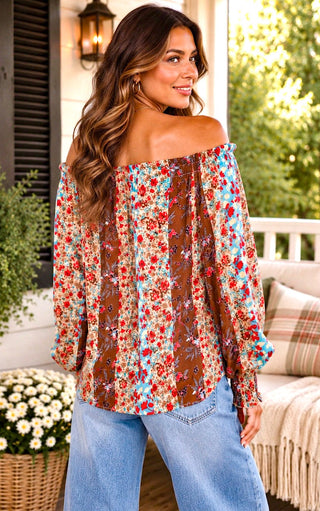 Back view of a woman wearing a brown off-the-shoulder blouse with a vibrant red, blue, and yellow floral pattern, paired with light blue jeans, standing on a porch.