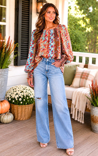 A woman stands on a porch wearing a long-sleeve bohemian floral print top and light-wash wide-leg jeans . The background features a white porch sofa with pillows, potted plants, and pumpkins.