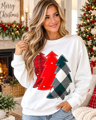 A woman wearing a white crewneck sweatshirt with a graphic of three Christmas trees in red, green, and black-and-white herringbone plaid patterns, posed in a cozy holiday living room setting.