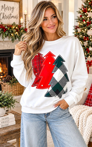 A woman wearing a white crewneck sweatshirt with a graphic of three Christmas trees in red, green, and black-and-white houndstooth patterns, posed in a cozy holiday living room setting.
