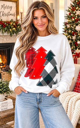 A woman wearing a white crewneck sweatshirt with a graphic of two Christmas trees in red and green plaid patterns, paired with light blue jeans, posed in a cozy holiday living room setting.