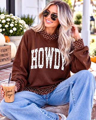 A woman wearing a brown crewneck sweatshirt with 'HOWDY' printed in white polka-dot letters across the chest, paired with light blue jeans and sunglasses, sitting outdoors and holding an iced coffee.