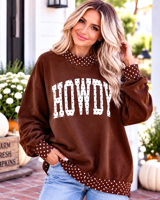 A woman wearing a brown crewneck sweatshirt with 'HOWDY' printed in white across the chest and white polka-dot trim on the collar, cuffs, and hem, paired with light blue jeans in an outdoor fall-themed setting.