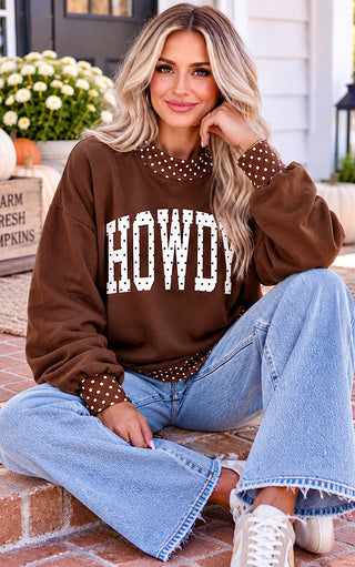 A woman sits on a brick porch wearing a brown crewneck sweatshirt with 'HOWDY' printed in white varsity letters across the chest, featuring brown and white polka-dot trim at the collar and cuffs. She is wearing light-wash blue jeans and white sneakers.