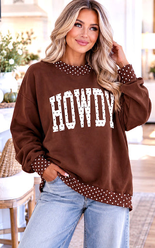 A woman wearing a brown crewneck sweatshirt with white polka-dot trim and large white 'HOWDY' lettering on the front, paired with light blue jeans in a bright indoor setting.
