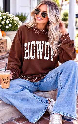 A woman wearing a brown crewneck sweatshirt with 'HOWDY' in white varsity letters and a polka-dot collar, paired with light blue jeans and sunglasses, sitting outdoors on a brick patio.