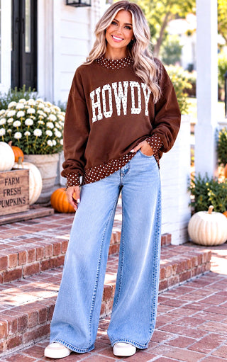 A woman stands on brick steps outdoors wearing a brown crewneck sweatshirt with 'HOWDY' in white varsity letters and a polka-dot mock neck and hem, paired with light blue wide-leg jeans and white sneakers.