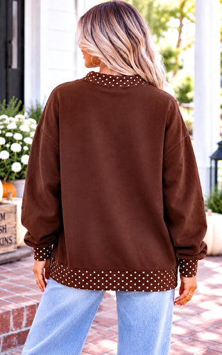 A woman viewed from behind wearing a brown long-sleeve sweatshirt with white polka dot trim on the collar and hem, paired with light blue jeans in an outdoor porch setting.