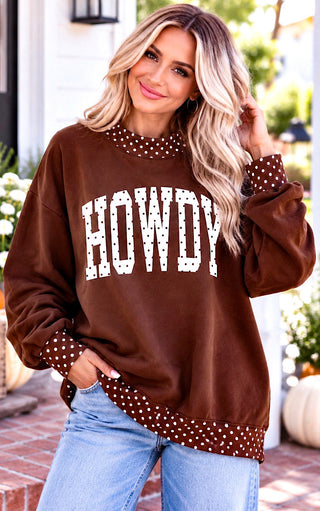 A woman wearing a brown crewneck sweatshirt with 'HOWDY' printed in white block letters with polka-dot trim, paired with light blue jeans, standing on a brick patio.