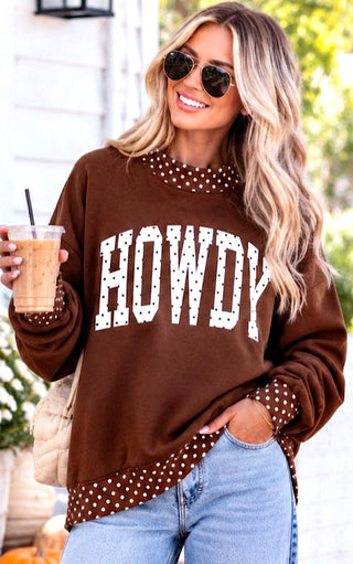 A woman wearing a brown crewneck sweatshirt with 'HOWDY' printed in white across the front, featuring white polka-dot trim at the collar, cuffs, and hem. She is wearing light blue jeans, sunglasses, and holding an iced coffee in an outdoor setting.