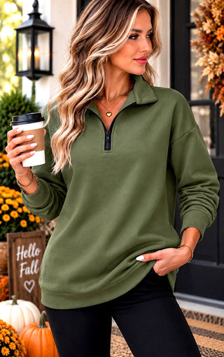 A woman wearing an olive green long-sleeve quarter-zip pullover sweatshirt, holding a coffee cup in a fall-themed outdoor setting with pumpkins and autumn decorations.