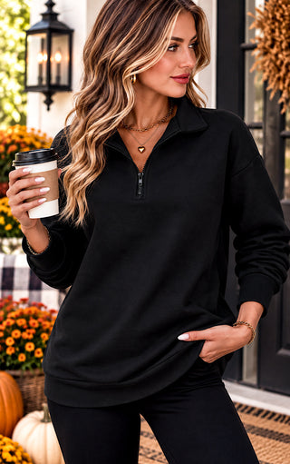A woman wearing a black long-sleeve quarter-zip pullover sweatshirt, holding a coffee cup in a fall-themed outdoor setting with pumpkins and flowers.