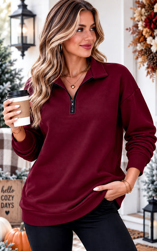 A woman wearing a long-sleeve quarter-zip pullover in deep burgundy, holding a coffee cup in a cozy, autumn-themed outdoor setting.