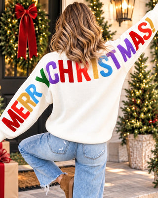A woman photographed from behind wearing a white sweater with the word 'MERRY' on the left sleeve and 'CHRISTMAS!' across the back in large rainbow-colored letters. She is standing outdoors in front of decorated Christmas trees and a lit doorway, wearing light blue jeans and brown boots.