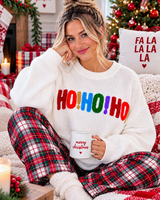 A woman sitting in a cozy holiday setting wearing a white knit sweater with 'HO! HO! HO!' in colorful letters and red/green plaid pajama pants, holding a white mug that says 'merry christmas'.