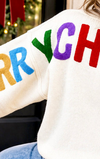A close-up of a person wearing a cream-colored fuzzy sweater with large colorful letters spelling 'MERRY CH.