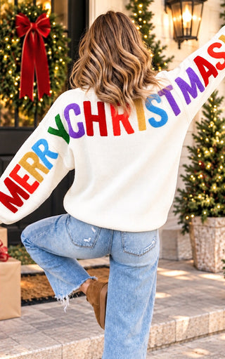 A woman seen from behind wearing a white crewneck sweater with 'MERRY CHRISTMAS' chenille letters across the back in rainbow colors, paired with light blue jeans, standing in front of a decorated Christmas tree and door.