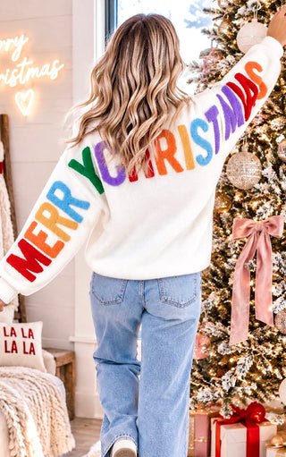 A woman viewed from behind wearing a white fuzzy sweater with 'MERRY CHRISTMAS' written across the back in colorful rainbow letters, standing next to a decorated Christmas tree in a cozy indoor setting.