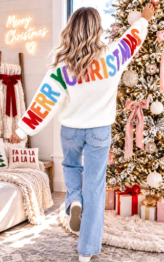 A woman viewed from behind wearing a white Christmas sweater with 'MERRY CHRISTMAS' printed across the back in rainbow colors, paired with light blue jeans, standing in a decorated living room next to a Christmas tree.
