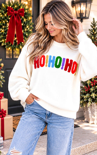 A woman wearing a cream-colored Christmas sweater with 'HO!HO!HO!' in colorful red, orange, yellow, green, blue, and purple letters, paired with light-wash blue jeans, set against a festive holiday background with wreaths and gifts.