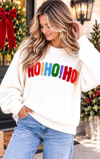 A woman wearing a cream-colored crewneck sweater with 'HO!HO!HO!' in colorful red, blue, green, and yellow letters, paired with blue jeans, set against a festive holiday background.