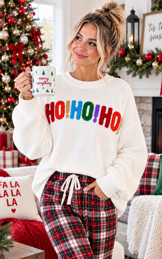 A woman in a cozy living room setting wearing a white crewneck sweater with 'HO! HO! HO!' in colorful letters and red plaid pajama pants, holding a mug that says 'merry christmas'.