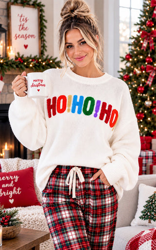 A woman wearing a white fuzzy crewneck sweater with 'HO! HO! HO!' in rainbow colors and red plaid pajama pants, holding a white mug that says 'merry christmas' in a cozy holiday living room.