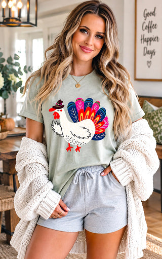 A woman wearing a sage green graphic t-shirt with a colorful turkey design, styled with a cream knit cardigan and light gray shorts in a cozy indoor setting.