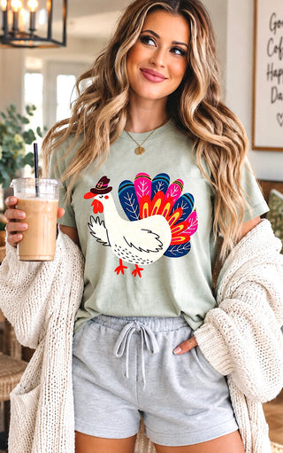 A woman wearing a sage green graphic t-shirt with a colorful turkey design, styled with light gray drawstring shorts and a cream knit cardigan, holding an iced coffee in a cozy indoor setting.