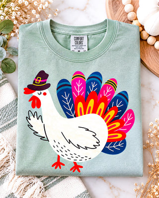 A folded sage green t-shirt featuring a colorful graphic of a turkey wearing a  Pilgrim hat, displayed on a marble surface with holiday-themed flat-lay props.