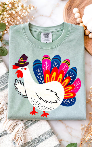 A folded sage green t-shirt with a colorful graphic of a white turkey wearing a black pilgrim hat, surrounded by autumn-themed flat-lay props like dried flowers, a striped blanket, and a wooden tray.