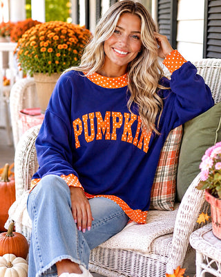 A woman sitting on a white wicker porch chair wearing a royal blue crewneck sweatshirt with 'PUMPKIN' written in orange polka-dot letters across the chest, with orange polka-dot trim on the collar and cuffs.