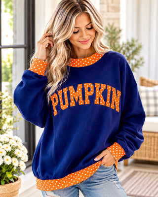 A woman wearing a navy blue fleece sweatshirt with 'PUMPKIN' printed in orange letters with white polka dots, and matching orange polka dot trim on the collar, cuffs, and hem, paired with light wash jeans.