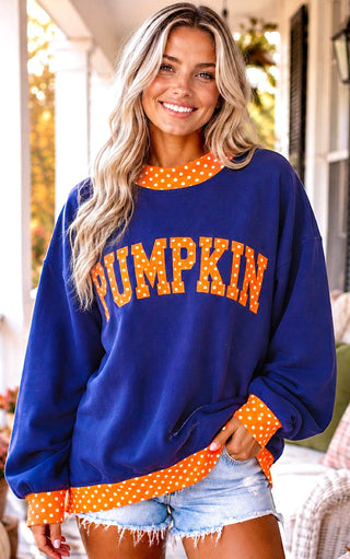 A woman wearing a royal blue crewneck sweatshirt with orange and white polka-dot trim and 'PUMPKIN' printed across the chest in orange letters, paired with denim shorts.