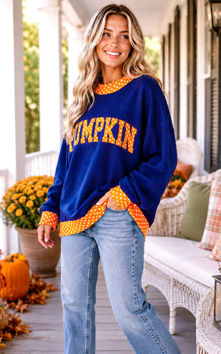 A woman stands on a porch wearing a navy blue crewneck sweatshirt with 'PUMPKIN' written across the front in orange letters with white polka dots, paired with light blue jeans. The background shows a cozy porch setting with pumpkins, mums, and a white wicker bench.