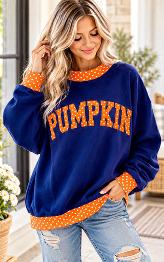 A woman wearing a navy blue sweatshirt with orange and white polka-dot trim and 'PUMPKIN' written across the front in orange polka-dot letters, paired with light blue ripped jeans.