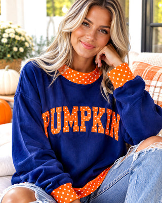 A woman wearing a navy blue sweatshirt with orange and white polka-dot trim and 'PUMPKIN' printed across the front in orange letters with white polka dots. She is sitting on a couch in a cozy home setting.