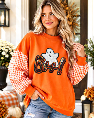 A woman wearing an orange crewneck sweatshirt with gingham checkered sleeves and a sequined 'Boo!' graphic, posing in front of a decorated autumn doorway.