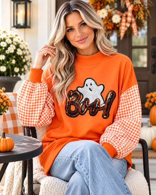 A woman wearing an orange Halloween sweatshirt with orange-and-white gingham sleeves and a sequined ghost and 'Boo!' graphic, seated outdoors on a porch with pumpkins and fall decorations in the background.