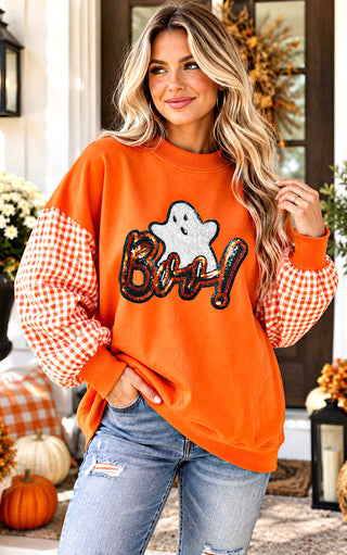 A woman wearing a bright orange crewneck sweatshirt with gingham checkered sleeves and a 'Boo!' graphic with a ghost on the front, paired with distressed blue jeans, posed in front of a decorated autumn doorway.