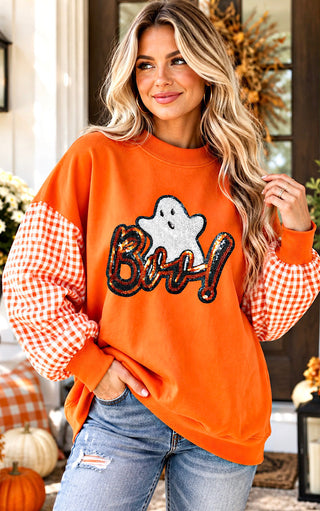 A woman wearing an orange crewneck sweatshirt with gingham checkered sleeves and a sequined 'BOO’ ghost graphic on the front, paired with distressed blue jeans in a fall-themed home setting.